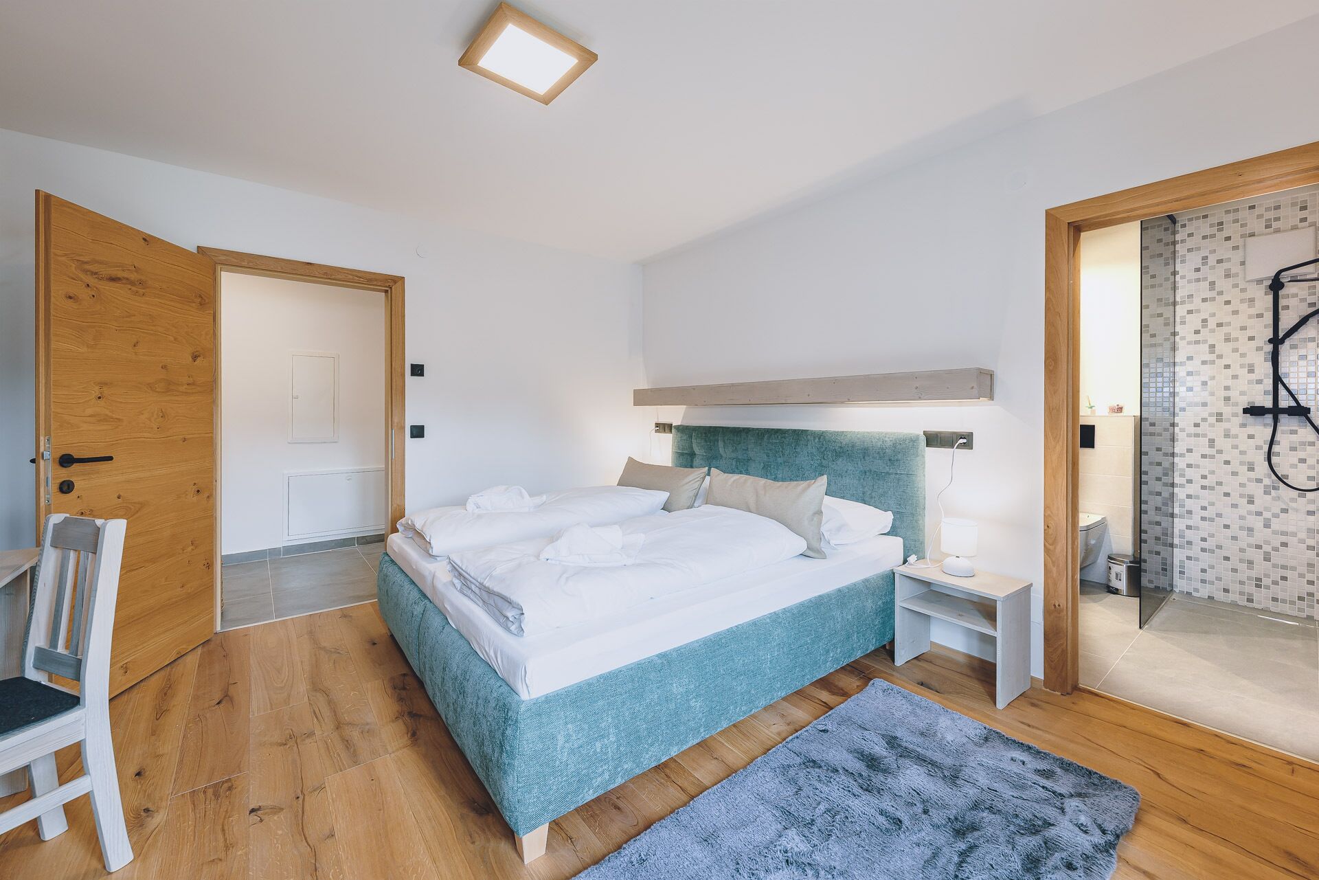A modern bedroom with a large bed, a bedside table, a chair, and a shower area visible through an open door.