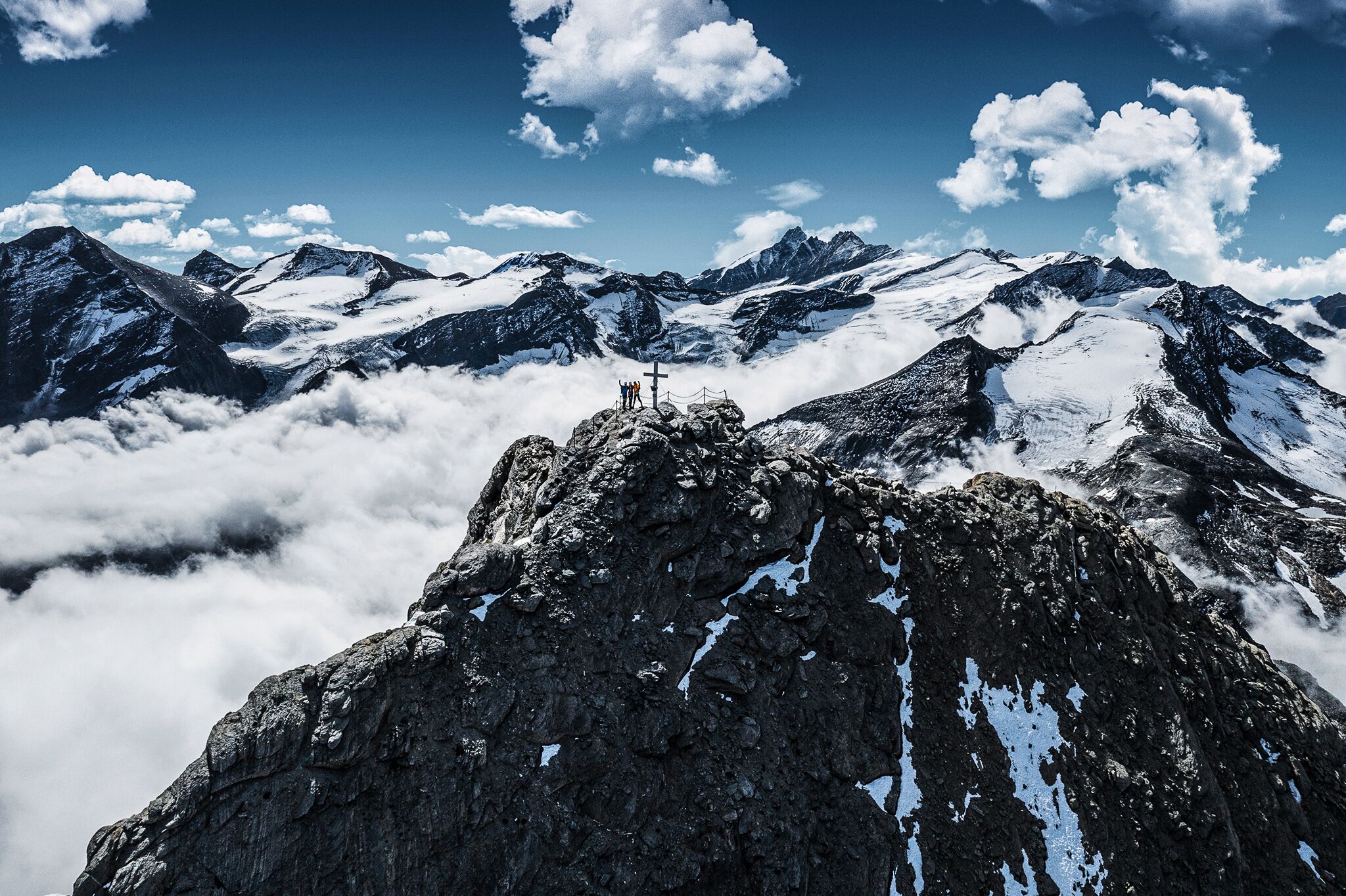 A mountain peak with a cross, surrounded by snow-covered peaks and clouds.