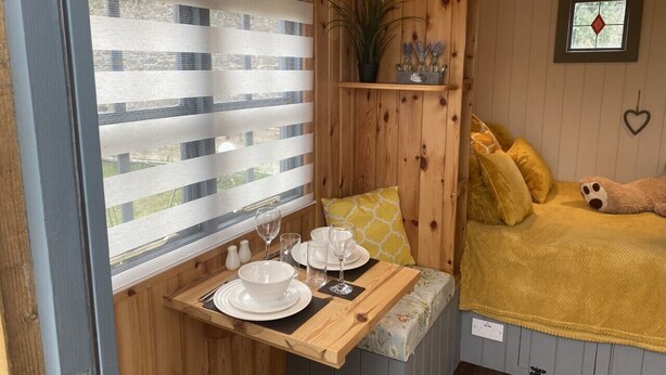 Una habitación acogedora con detalles de madera, una cama con manta amarilla, una mesita con platos y vasos blancos, una ventana con persianas a rayas y un estante con adornos.