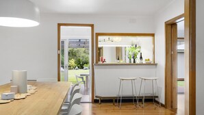Een moderne eetruimte met een houten tafel, witte stoelen en een bank. Er is een keukenblad met een spiegel erboven en uitzicht op een tuin door een geopende deur.