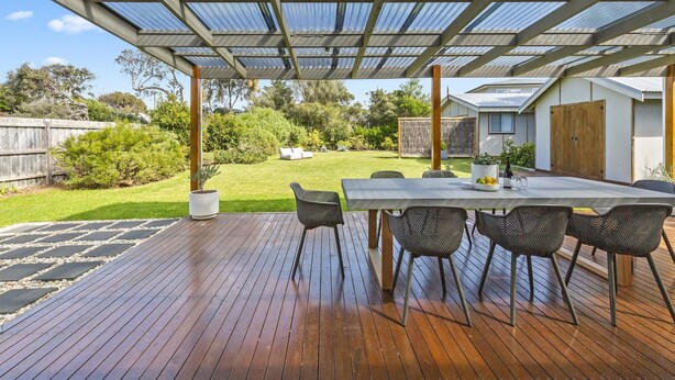 Een overdekt terras met een houten terras, een tafel met stoelen en een uitzicht op een huis op de achtergrond.