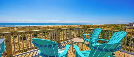 A wooden deck with turquoise Adirondack chairs and a small round table, overlooking a beach with dunes and ocean.