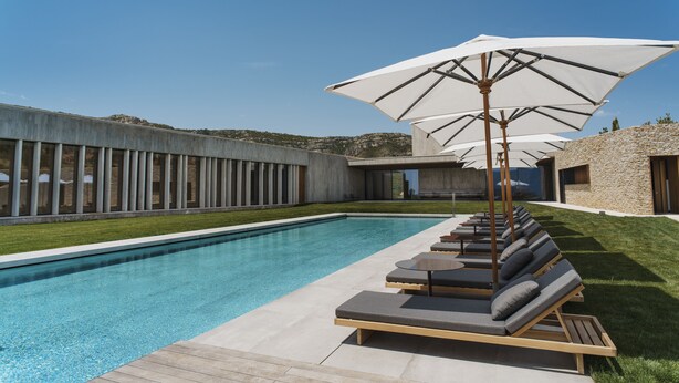 A modern outdoor pool area with lounge chairs and umbrellas, surrounded by stone buildings and a clear sky.