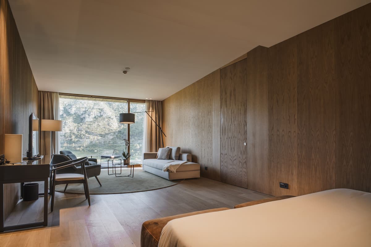 A modern hotel room with a large bed, a desk with a lamp, a sofa, and a view of the outdoors.