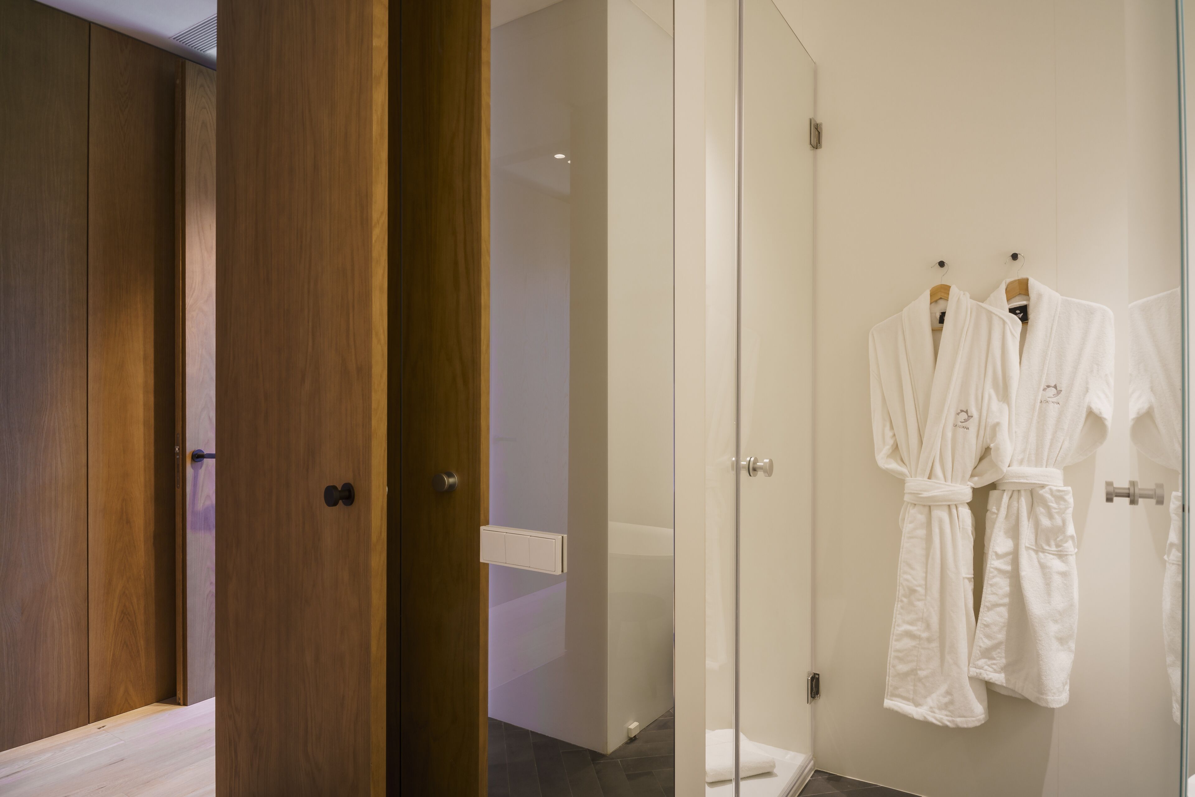 A modern bathroom with a glass shower enclosure, wooden wardrobe, and two white robes hanging on hooks.