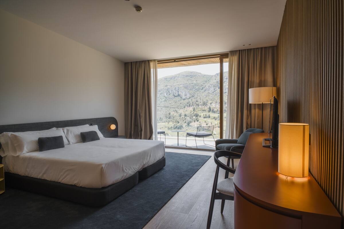A hotel room with a large bed, a desk with a lamp, a chair, and a view of mountains through the window.