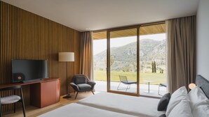 A modern hotel room with a large bed, a desk with a TV, a chair, and a view of a mountainous landscape through large windows.