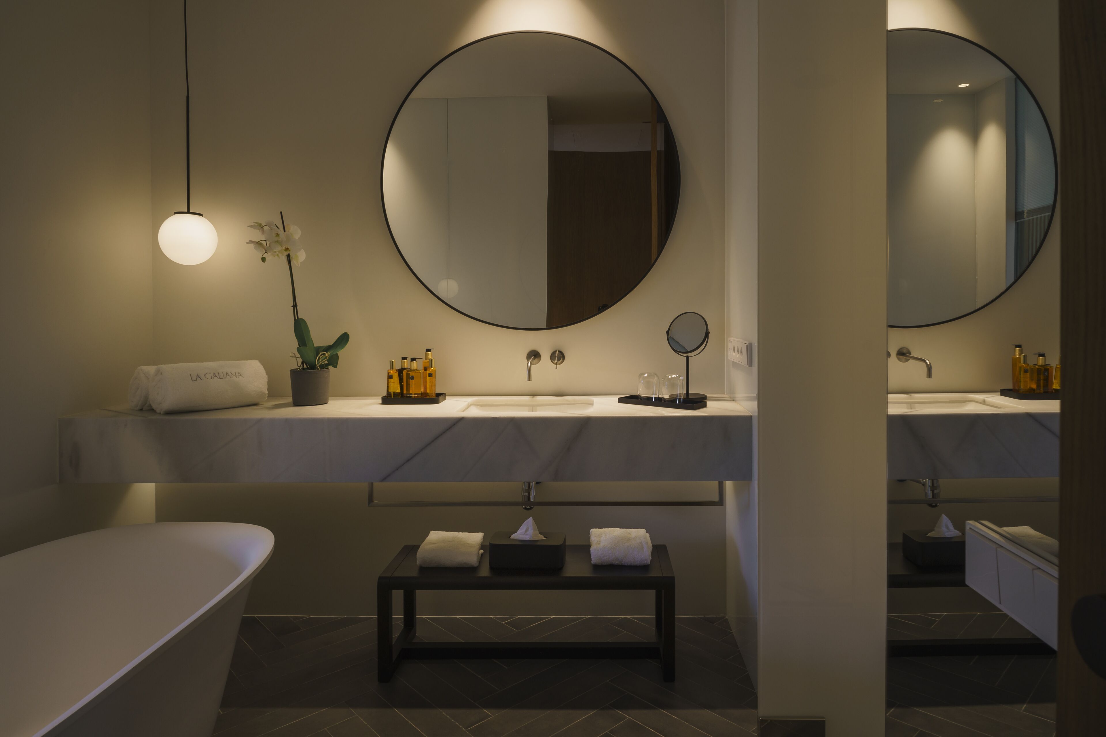 Ein modernes Badezimmer mit einem runden Spiegel, einer Badewanne, einer Sitzbank und einem Blumentopf.
