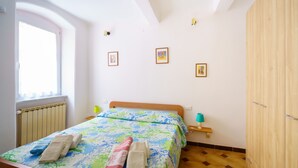 A bedroom with a bed, a window, a radiator, a wooden wardrobe, and a small bedside table with a lamp.