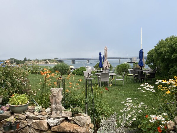 Ett utomhusområde med sittplatser, bord och stolar, en stenskulptur och utsikt över en bro som spänner över en vattenyta.