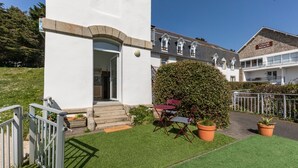 A white building with a stone facade, a small outdoor seating area with a table and chairs, and a neatly trimmed hedge. - La Tour du Castel Beach - 200m de la plage (Moëlan-sur-Mer)