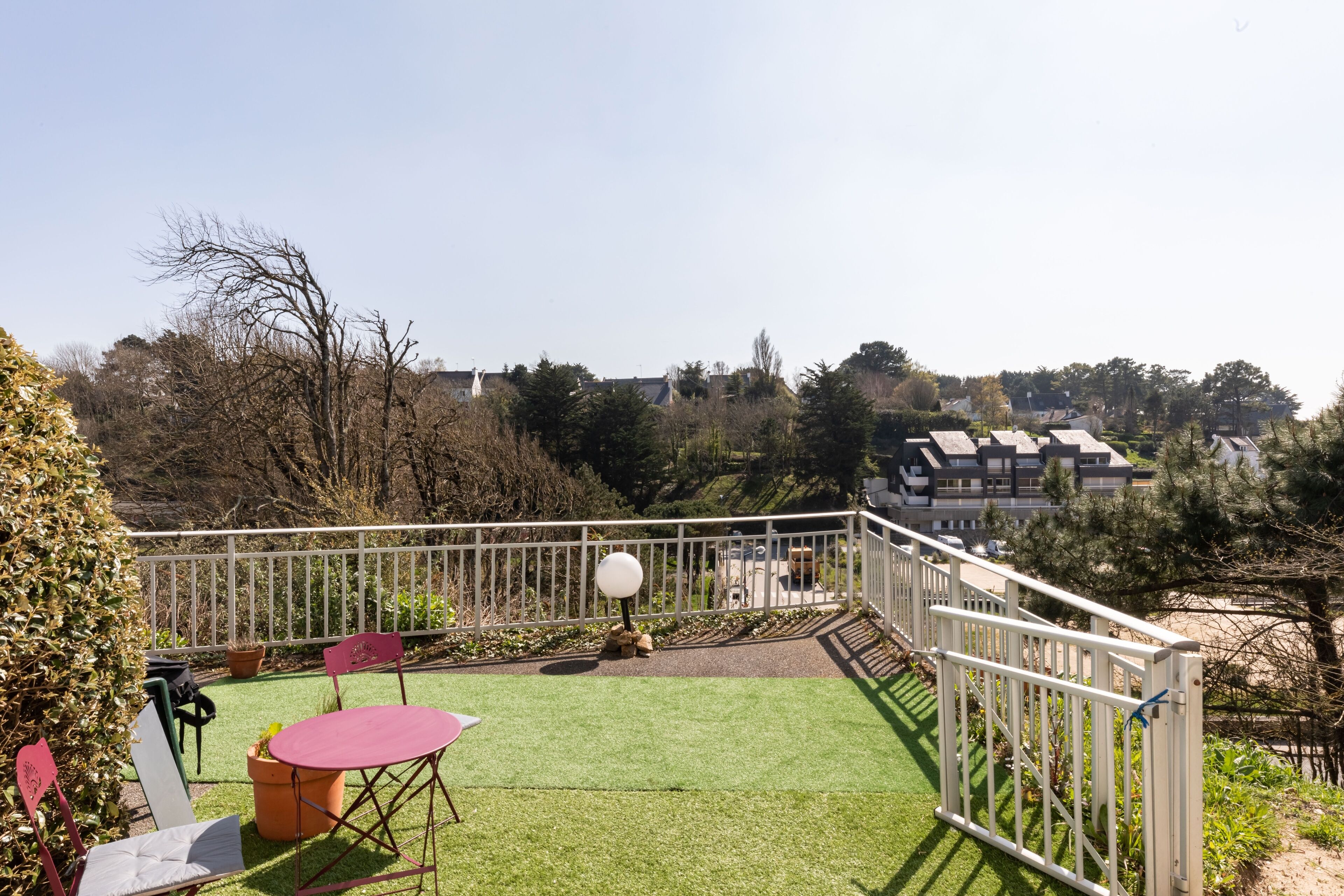 A balcony with a green lawn, a white railing, and a view of buildings in the distance.