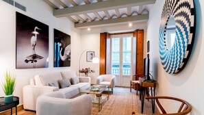 A modern living room with a sofa, armchairs, a coffee table, and a dining area with chairs. There are two large wall art pieces, a potted plant, and a mirror.