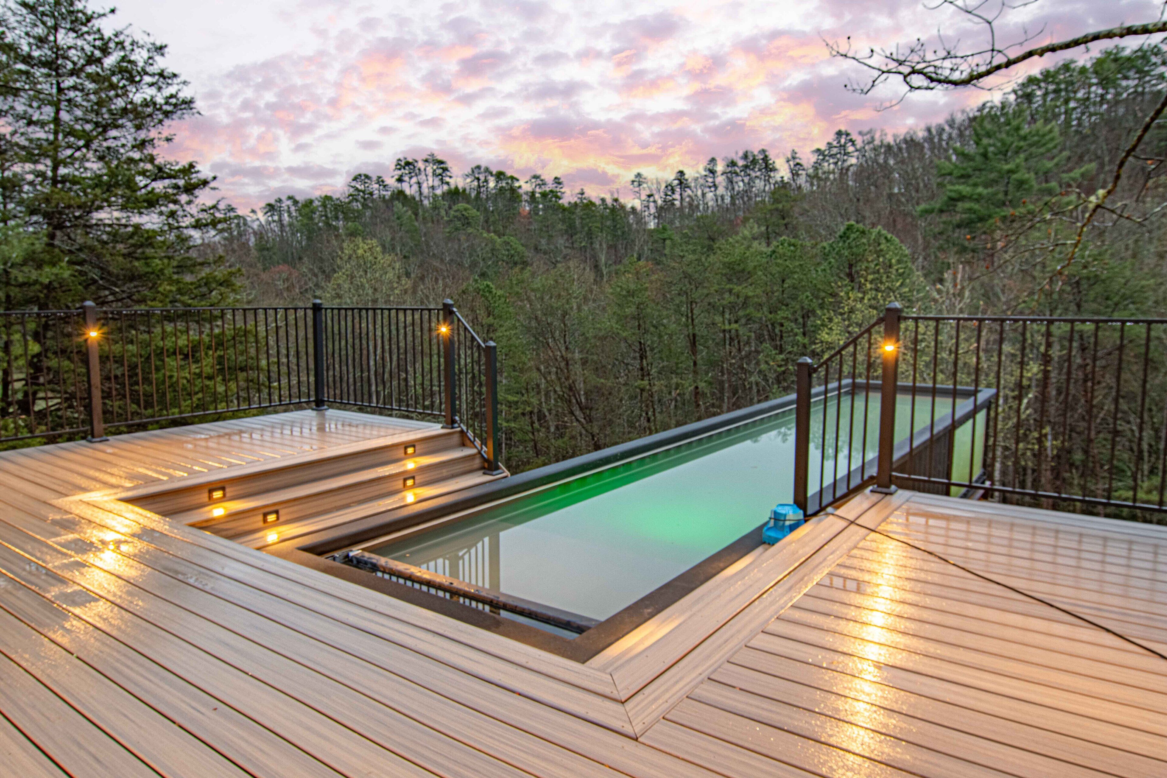 Eine Holzterrasse mit Schwimmbecken, umgeben von einem Metallgeländer und Bäumen.