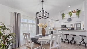 A modern kitchen with white cabinets, a stainless steel refrigerator, and a microwave. There is a dining area with a white table and chairs, and a hanging plant above the kitchen counter.