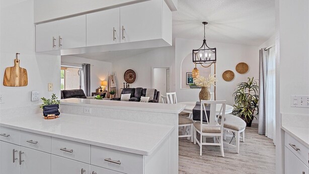 A modern kitchen with white cabinetry and a central island, a dining area with white chairs, and a living room with a sofa and coffee table.