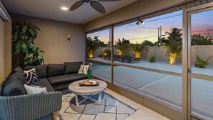 A modern living room with a sofa, coffee table, and a ceiling fan. Large windows offer a view of the outside.