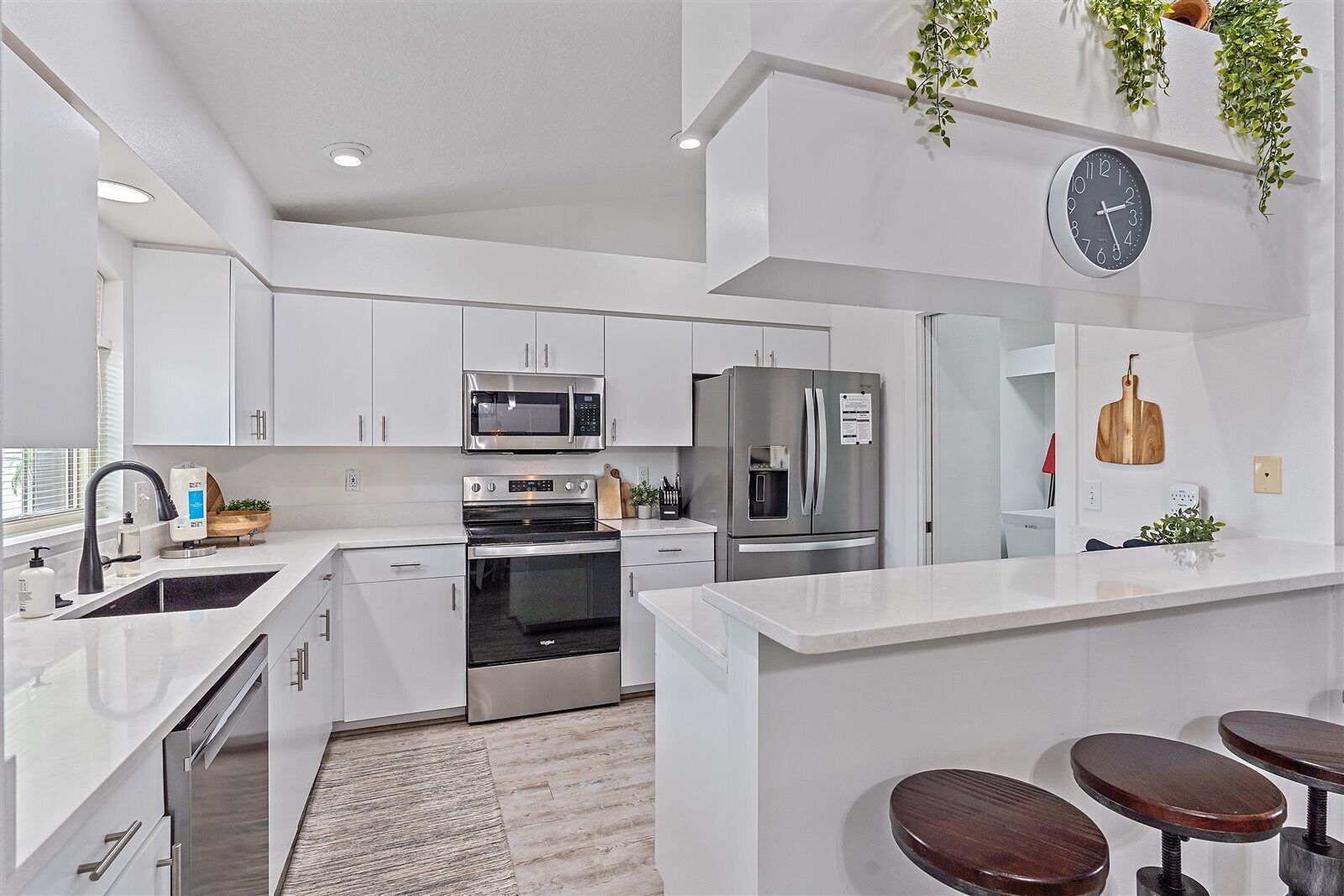 A modern kitchen with white cabinetry, stainless steel appliances, a built-in oven, a microwave, and a refrigerator. There is a clock on the wall and a plant hanging above the counter.