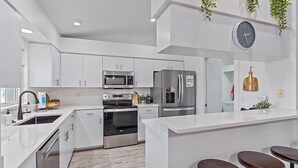 A modern kitchen with white cabinetry, stainless steel appliances, a built-in oven, a microwave, and a refrigerator. There is a clock on the wall and a plant hanging above the counter.