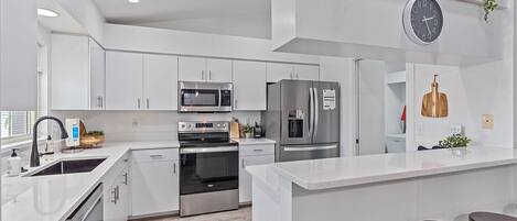A modern kitchen with white cabinetry, stainless steel appliances, a built-in oven, a microwave, and a refrigerator. There is a clock on the wall and a plant hanging above the counter.