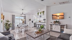 A modern living room with a dining area, kitchen, and a large TV mounted on the wall.