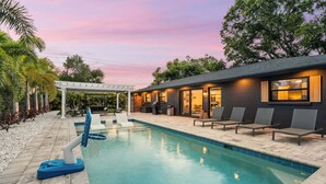 A swimming pool with lounge chairs, a pergola, and a building with large windows. - Sea Esta | Private Saltwater POOL + Putting Green! (Sarasota)
