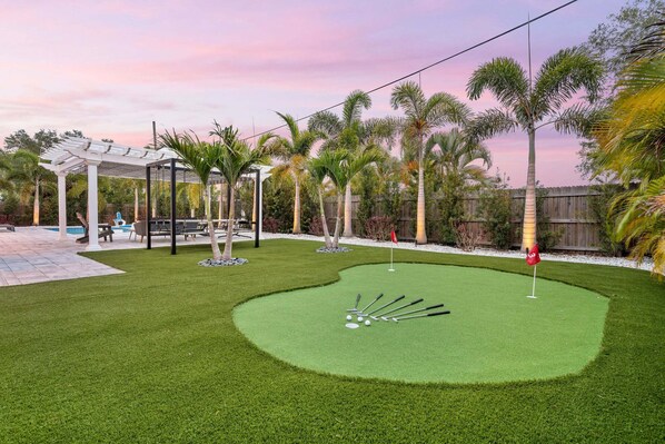 A backyard with a putting green, palm trees, a pergola, and a pool.