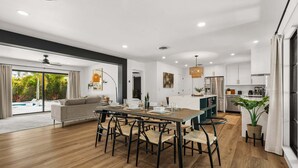 A modern kitchen with white cabinets, a dining area with a wooden table and chairs, and a large window with a view of a pool. - Sea Esta | Private Saltwater POOL + Putting Green! (Sarasota)