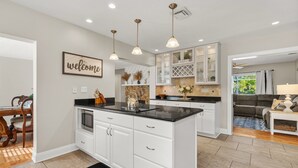 Una cocina moderna con armarios blancos, encimeras negras e iluminación colgante. Hay una zona de comedor con una mesa y sillas de madera, y una sala de estar con un sofá y una mesa de centro.