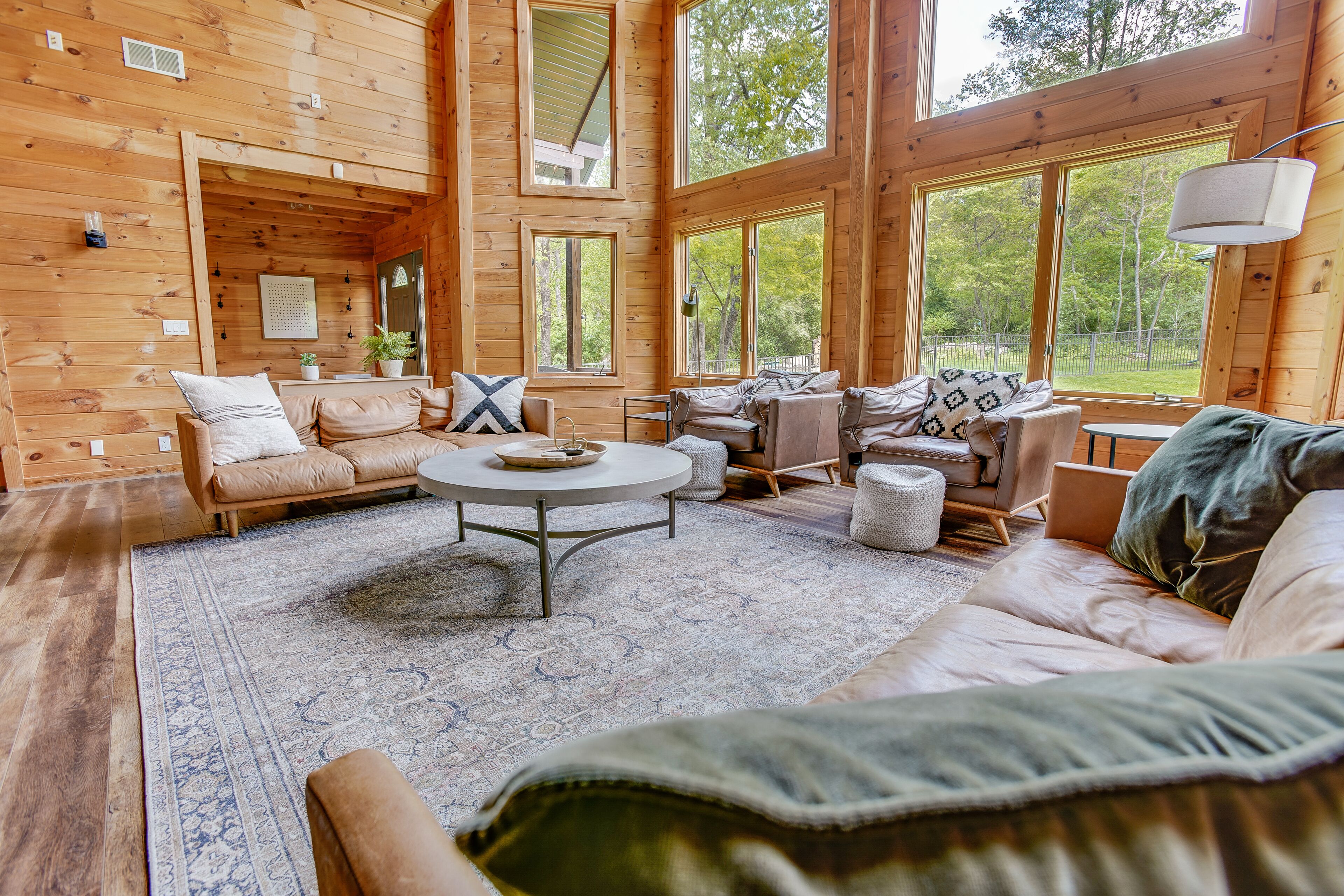 A spacious wooden cabin interior with a large living area, multiple seating options, and large windows offering views of the surrounding forest.