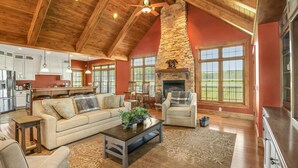 A spacious living area with a high ceiling, stone fireplace, and large windows offering views of the outdoors. - Kountry Lane Stables | Horse Farm w/ HOT TUB + Fire Pit! (Wakarusa)