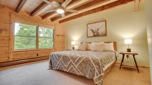Une chambre avec un plafond en bois, une grande fenêtre, un lit recouvert d’une couverture à motifs, deux lampes de chevet et un tableau encadré au mur.