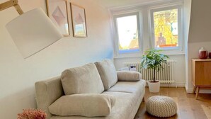 Un salon avec un canapé beige, une table d’appoint blanche ornée d’un vase, une plante en pot et un radiateur.