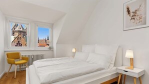 Une chambre à coucher comprenant un lit, deux tables de chevet, une chaise et une fenêtre donnant sur un immeuble.