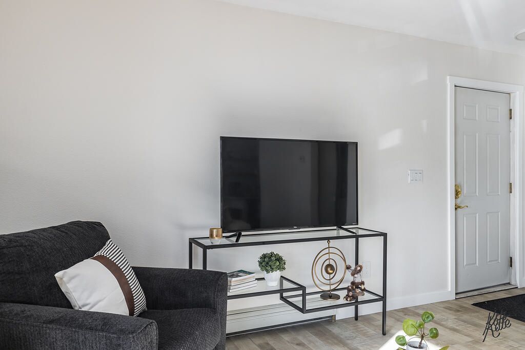 A modern living room with a flat-screen TV on a glass TV stand, a dark grey sofa with striped pillows, and a white door in the background.