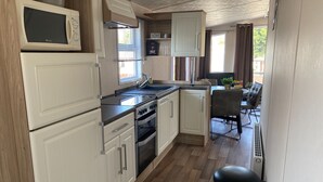 Cocina compacta con muebles blancos, microondas, estufa y lavaplatos. Se ve un área de comedor con mesa y sillas a través de una ventana.