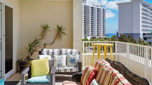 A balcony with wicker furniture, a yellow table, and a view of tall buildings and the ocean.