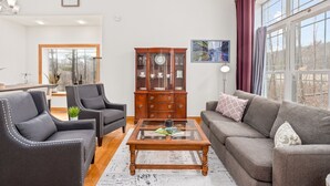 Un salon comprenant un canapé, deux fauteuils, une table basse en verre, un meuble en bois et une grande fenêtre avec des rideaux.