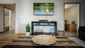 Un salon avec un téléviseur fixé au mur, une console en bois agrémentée d’objets décoratifs, un panier en osier posé sur un tapis et une porte donnant sur une autre pièce.