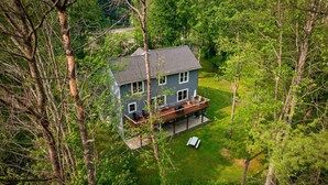 Une maison à deux étages dotée d’une terrasse, entourée d’arbres denses et d’une pelouse bien entretenue.