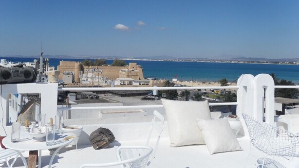 Une terrasse sur le toit, avec des meubles blancs, une table avec des verres et une bouteille, et une vue sur la mer et les bâtiments éloignés.