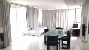 Une chambre d’hôtel avec une table à manger en verre, des chaises noires, un lit, un bureau avec un ordinateur et un téléviseur.
