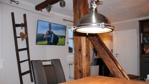 Zona pranzo con tavolo in legno, una sedia, televisore fissato alla parete e struttura con travi in legno.