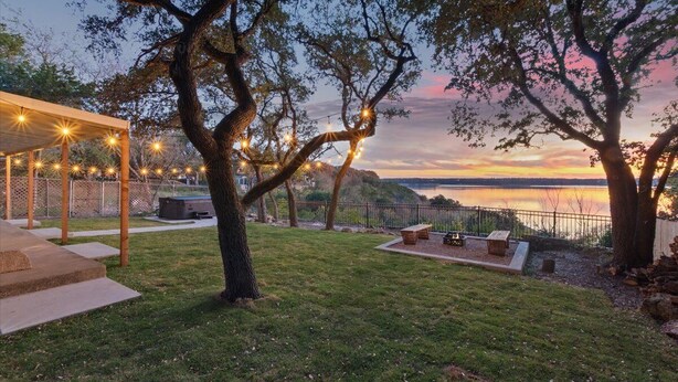 A backyard with a pergola, string lights, a fire pit, and a view of a lake at sunset.