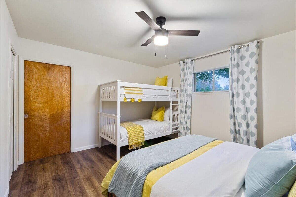 A bedroom with a bunk bed, a ceiling fan, a window with curtains, and a wooden door.