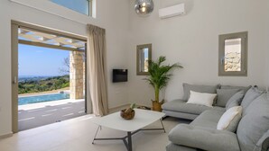 A modern living room with a grey sofa, a white coffee table, and a large potted plant. There is a TV mounted on the wall and a view of a swimming pool through an open door.