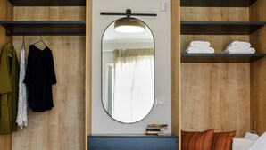 A bedroom with a wooden wardrobe, a mirror, a bedside table with books, a bed with pillows, and a shelf with folded towels.