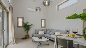 A modern living room with a grey sofa, a white coffee table, and a dining table with chairs. There are two windows with curtains, a potted plant, and a hanging light fixture.