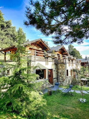 Un chalet rustique aux murs de pierre, doté de balcons en bois et d’une cour verdoyante avec des chaises longues.