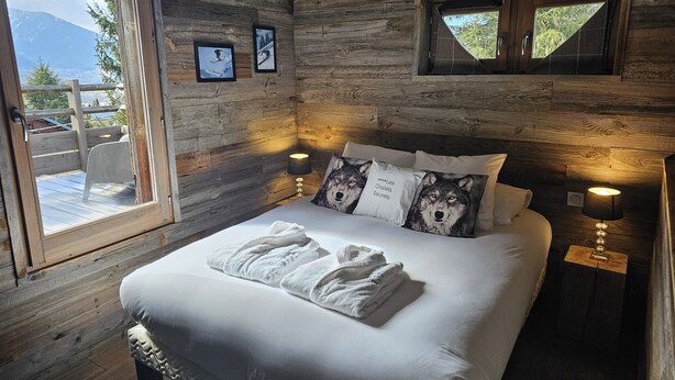 A wooden cabin bedroom with a bed, two pillows with wolf prints, a window, and a door leading to an outdoor area. - Chalet Ankaa Collection Greniers d'Alpages (BOLQUÈRE FONT-ROMEU)